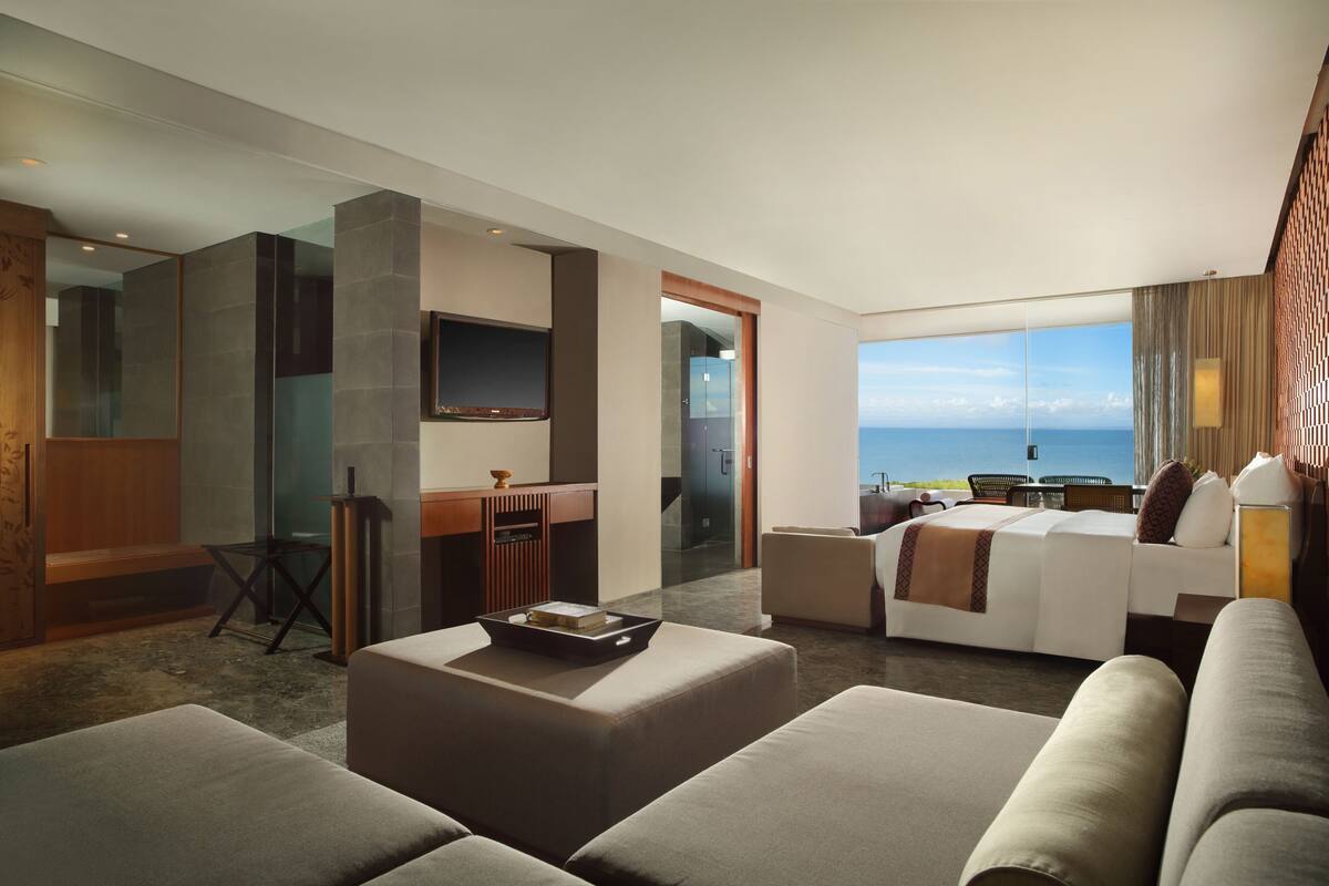 A modern hotel room with a large bed, a seating area with a coffee table, a TV mounted on the wall, and a view of the ocean.