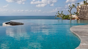 A clear blue pool with a curved edge, a rocky outcrop in the water, and a view of the ocean and sky with scattered clouds. - Anantara Uluwatu Bali Resort (Pecatu)