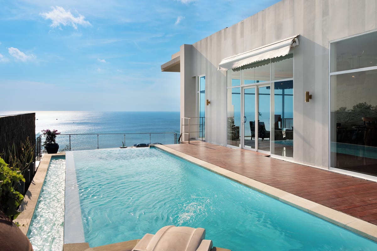 A modern poolside area with a view of the sea, a glass-enclosed patio, and a wooden deck.
