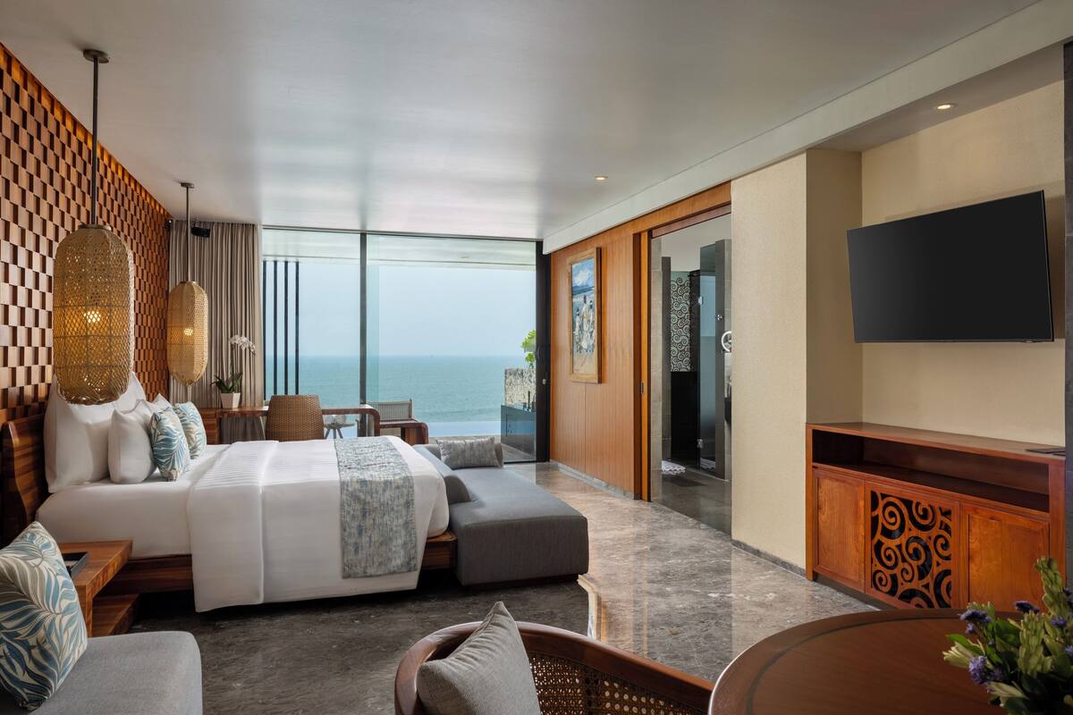 A modern hotel room with a large bed, a seating area, a TV, and a view of the ocean.
