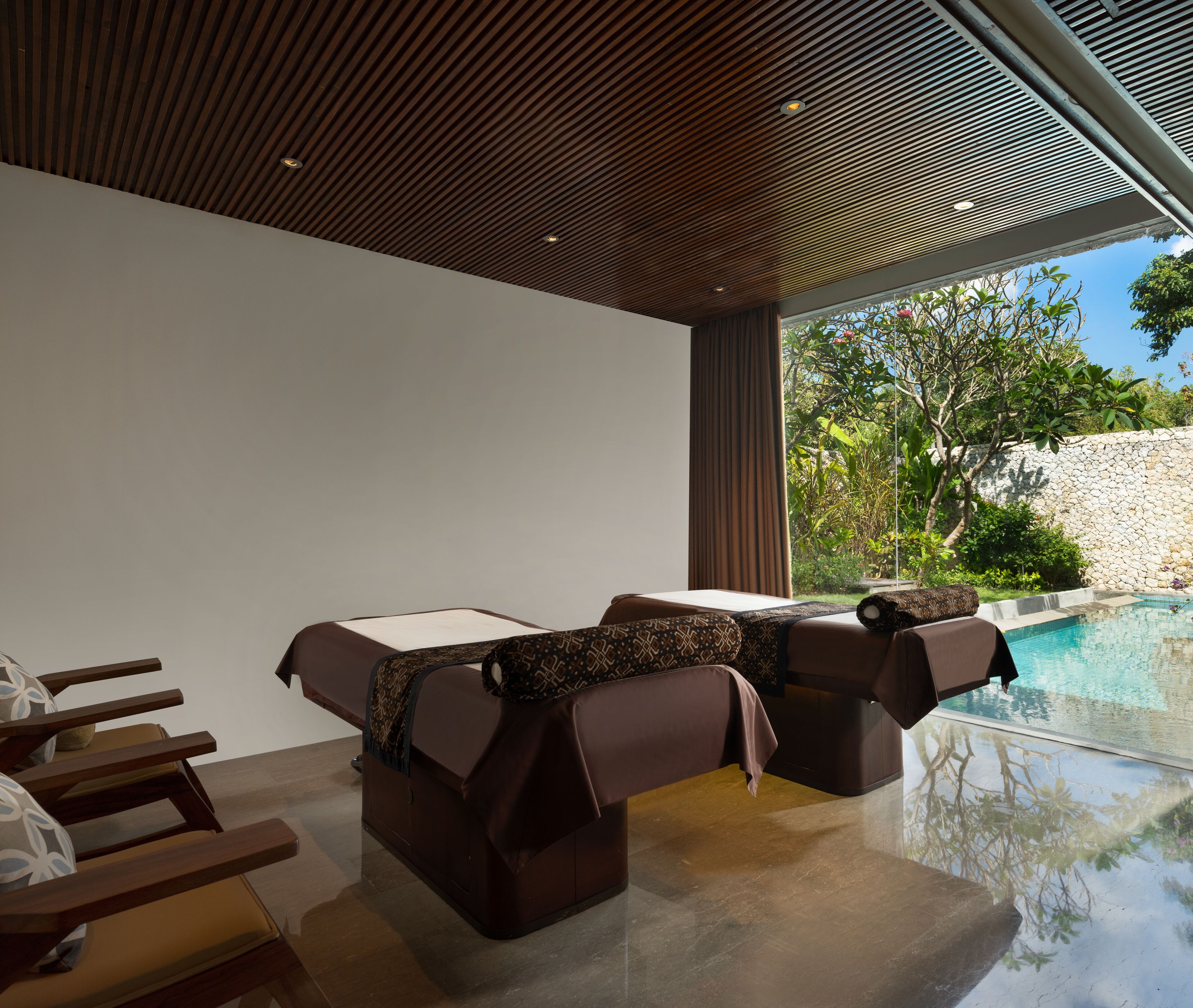 A modern lounge area with two massage chairs, a small table, and a view of a swimming pool.