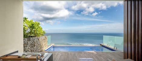 A balcony with a view of the ocean, a wooden deck, a bathtub, and a seating area with towels.