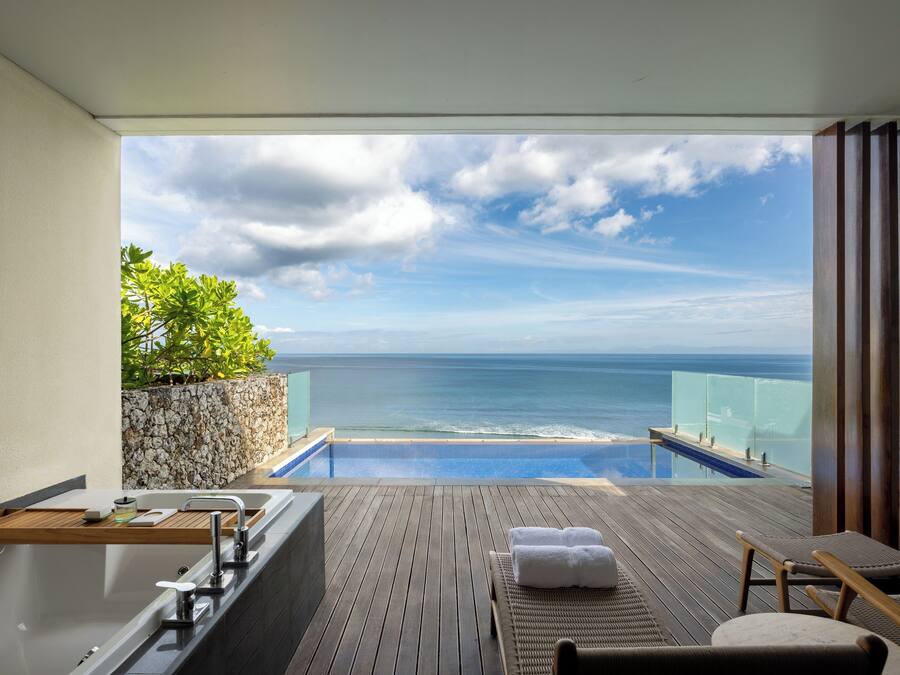 A balcony with a view of the ocean, a wooden deck, a bathtub, and a seating area with towels.