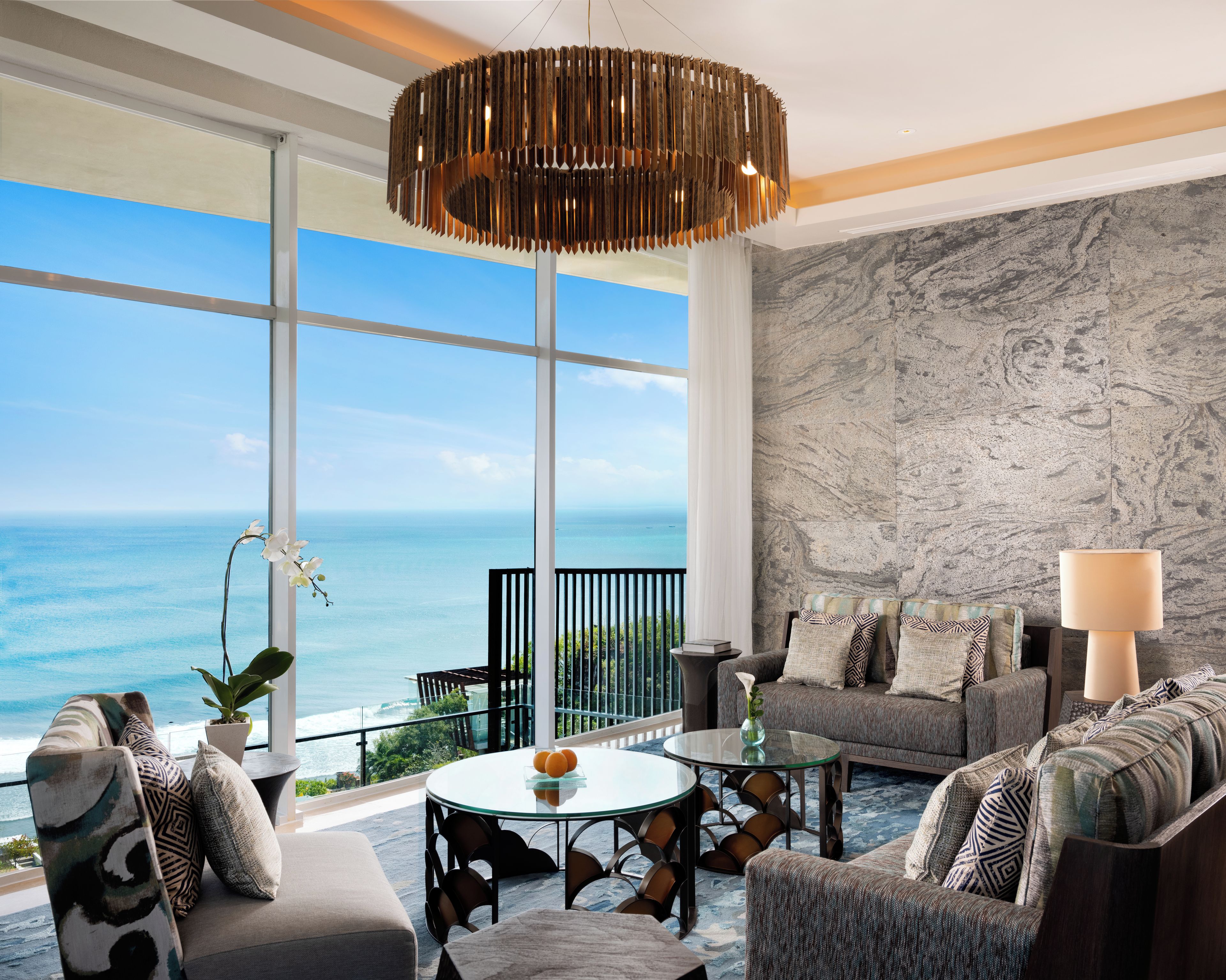 A modern living room with a view of the ocean, featuring a sofa, armchairs, a glass coffee table, and a unique hanging light fixture.