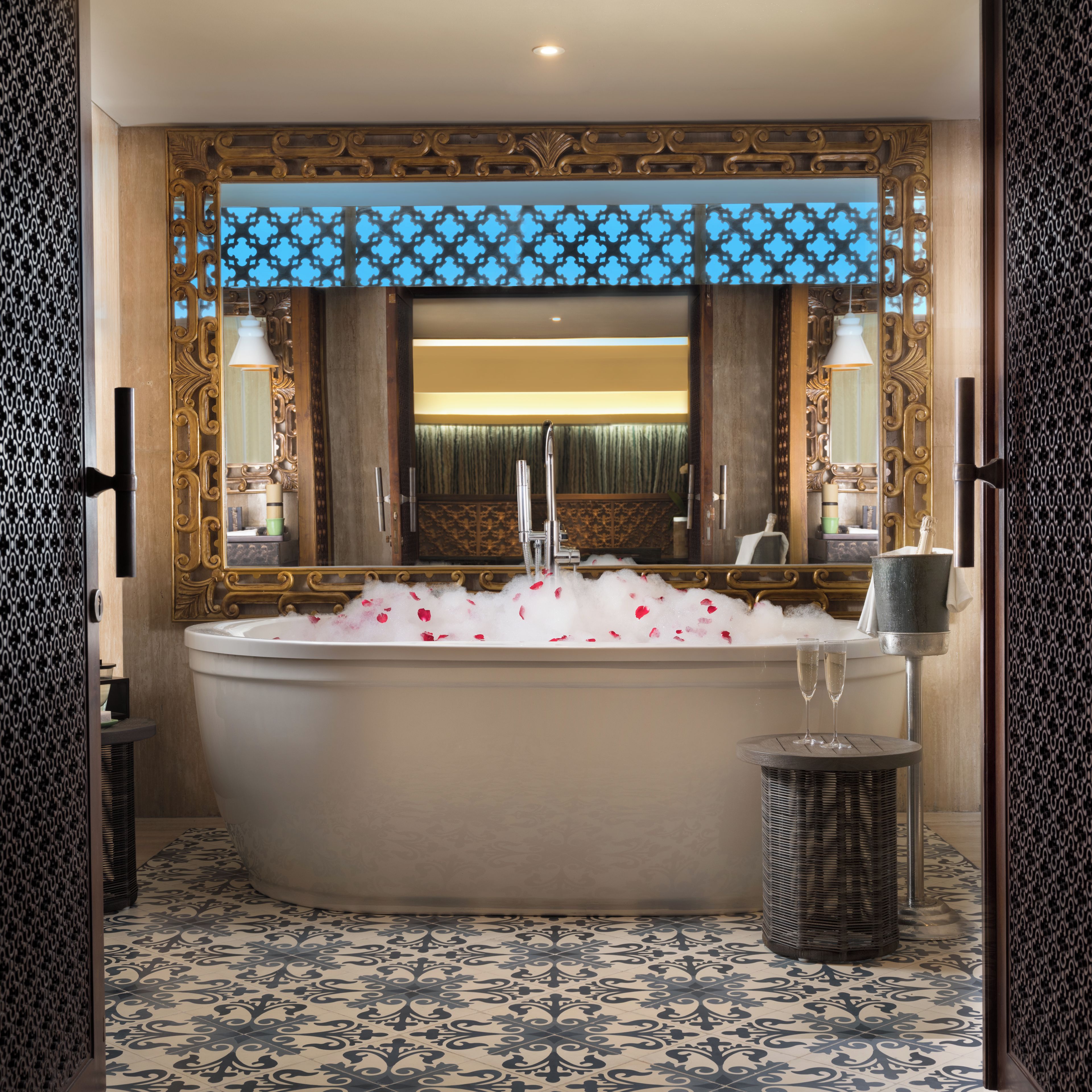 Un baño con una bañera independiente llena de pétalos de rosa, un espejo con detalles ornamentales dorados y un suelo estampado.