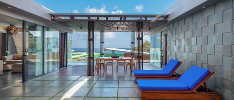 A modern outdoor lounge area with a glass enclosure, a pool table, and two blue lounge chairs.