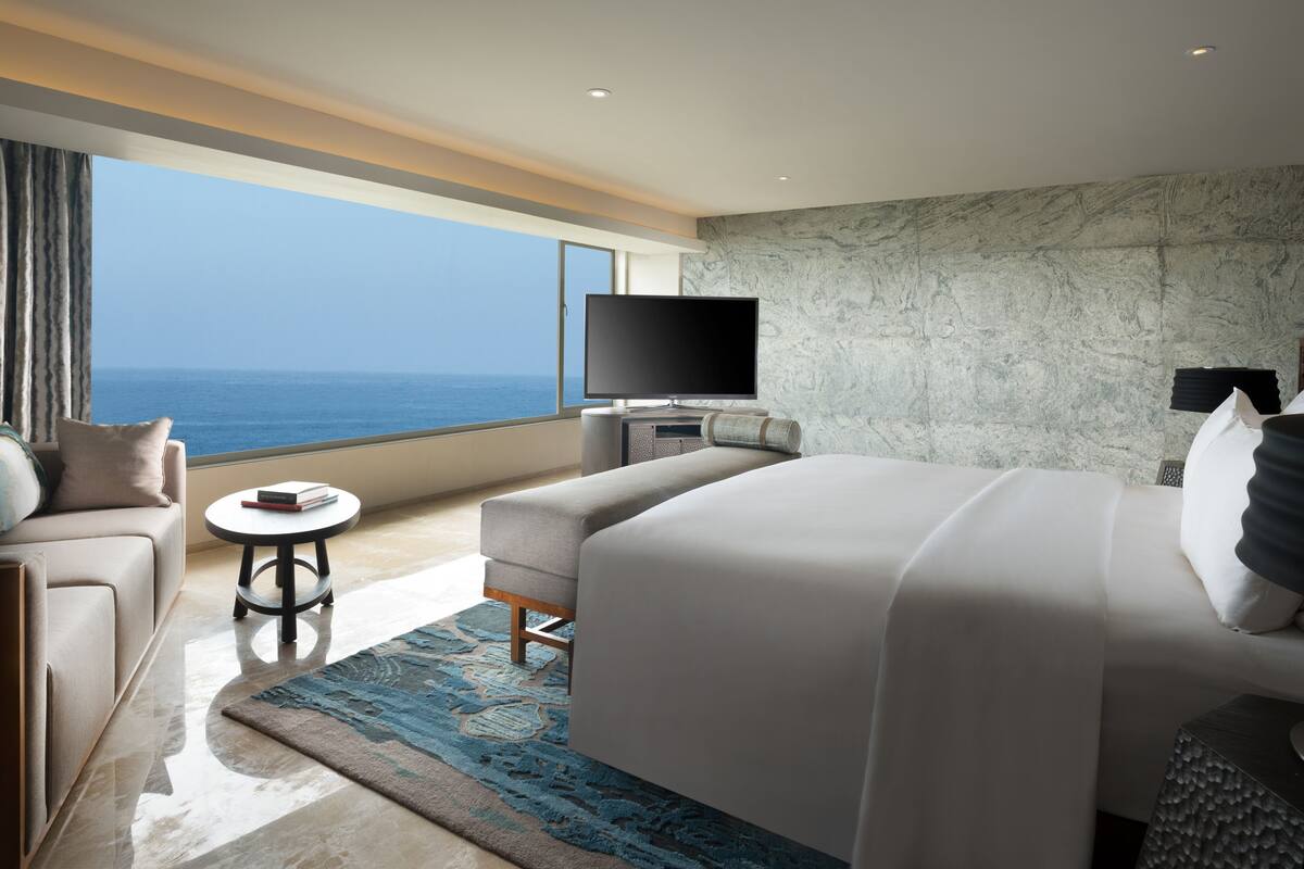 A modern hotel room with a large bed, a sofa, a small table, a flat-screen TV, and a view of the ocean.