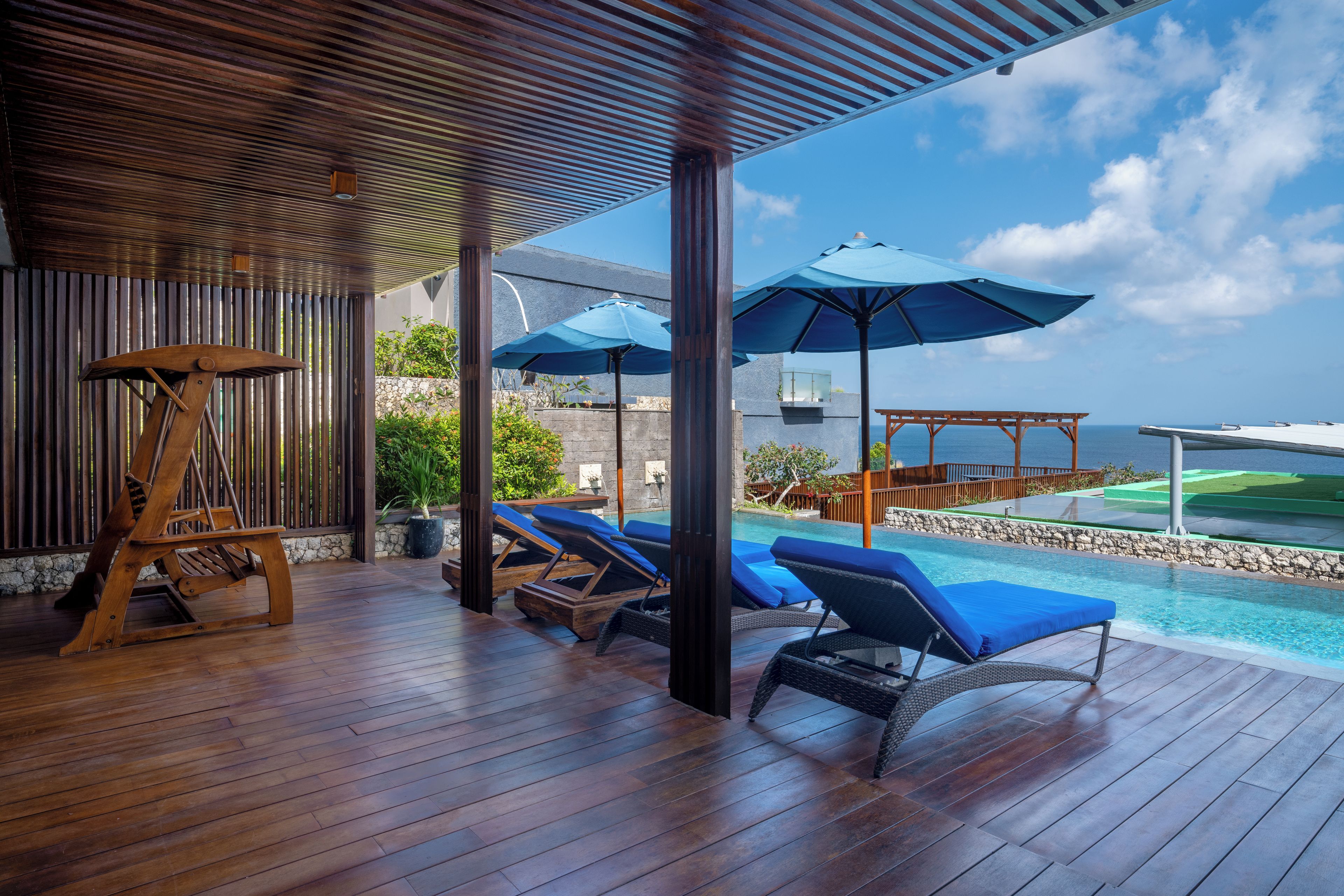 Terraza de madera con sillones de descanso y sombrillas, con vista a una piscina y al océano.