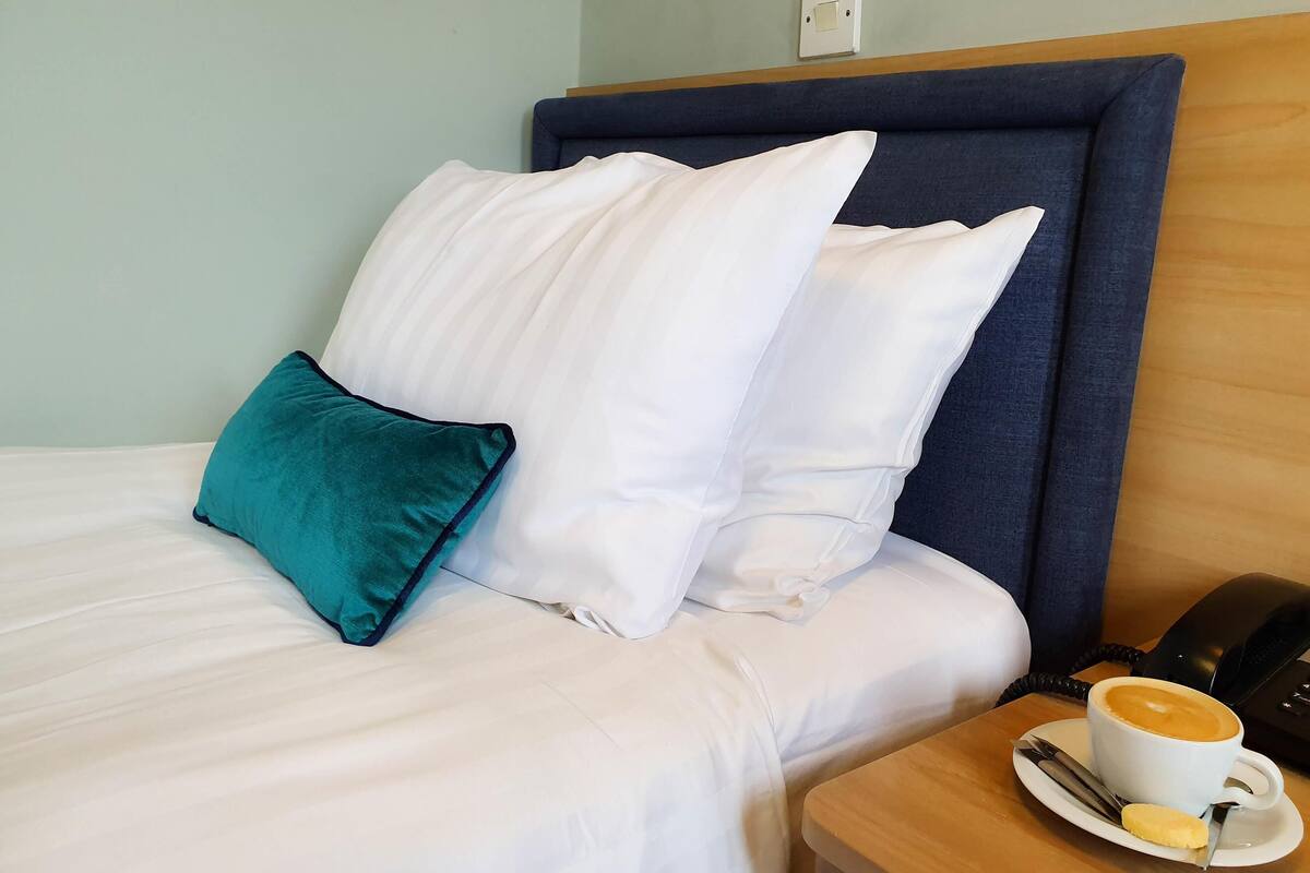 A hotel room with a bed, pillows, a bedside table with a coffee cup, and a telephone.