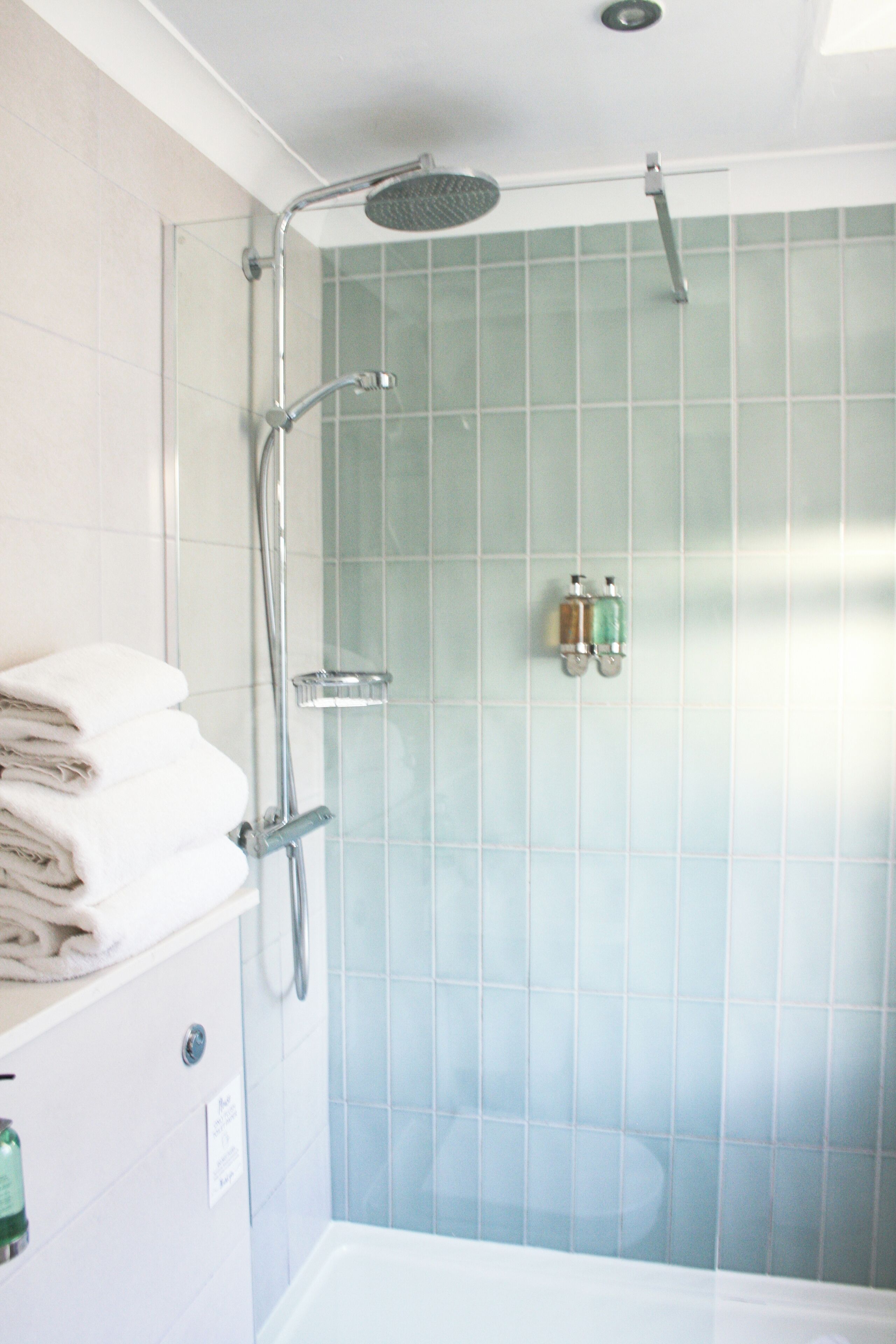 A bathroom with a glass-enclosed shower, white tiles, and a stack of white towels on a shelf.