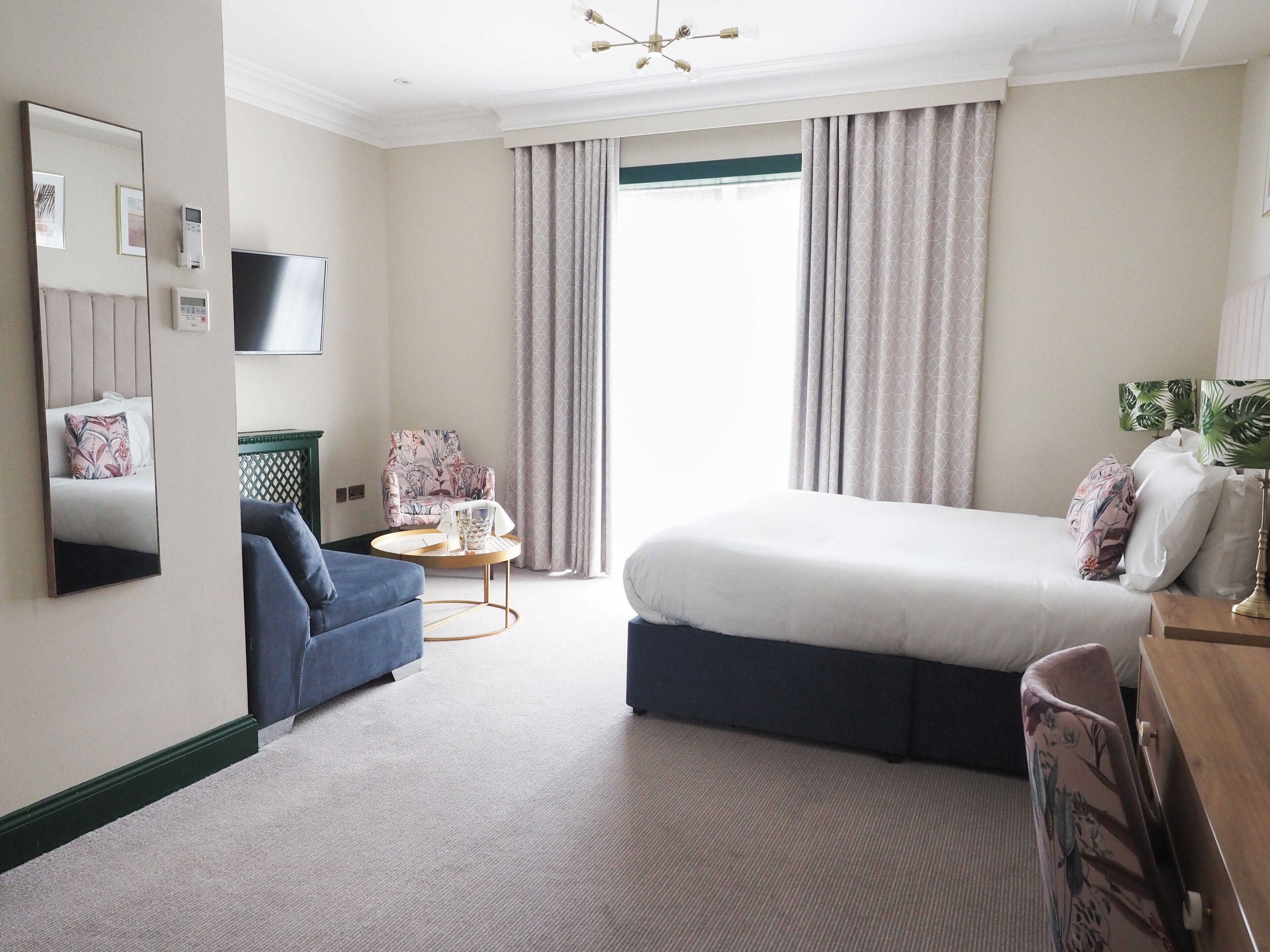 A hotel room with a bed, a blue armchair, a small round table, a TV, and a large mirror.