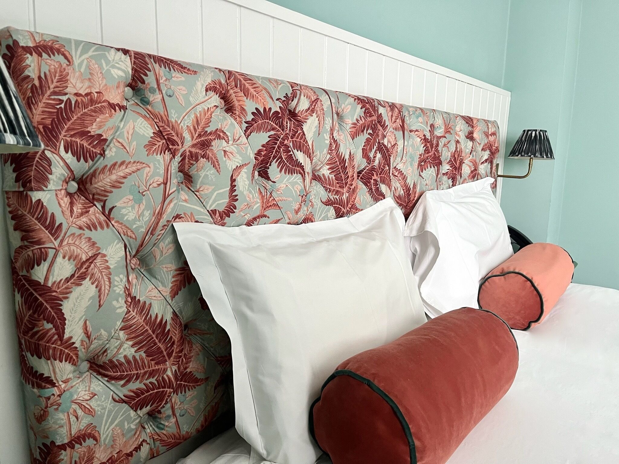 A bed with white pillows and a red accent pillow, a headboard with a floral pattern, and a lamp on the nightstand.