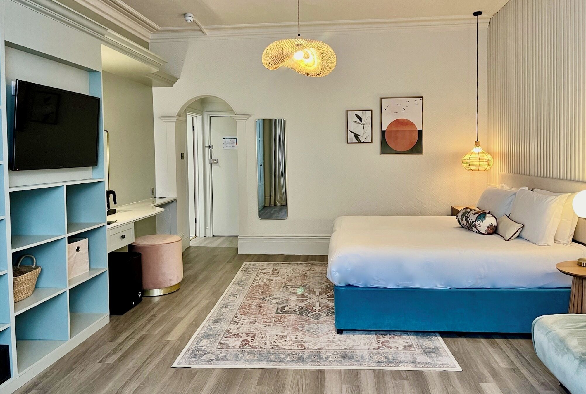 A bedroom with a bed, a TV, a wardrobe, a hanging light fixture, and a door leading to another room.