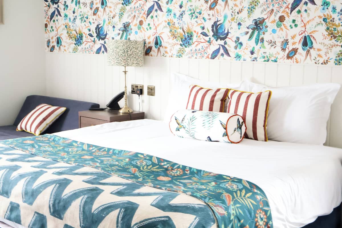 A neatly made bed with a floral-patterned headboard, a bedside table with a lamp, and a blue sofa with striped cushions.