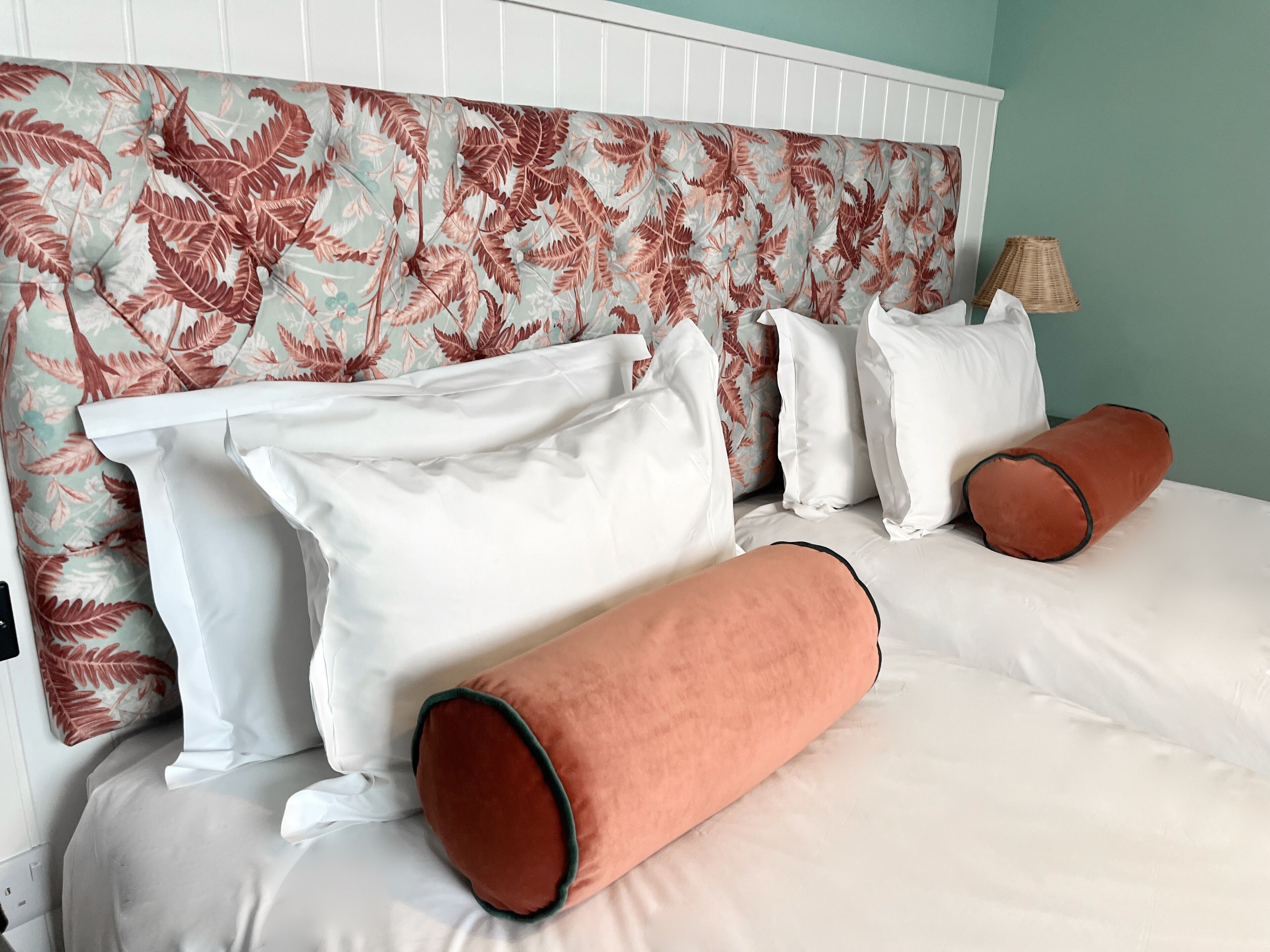 A neatly made bed with white linens and two orange pillows, a white headboard with a leafy pattern, and a lamp on the side.