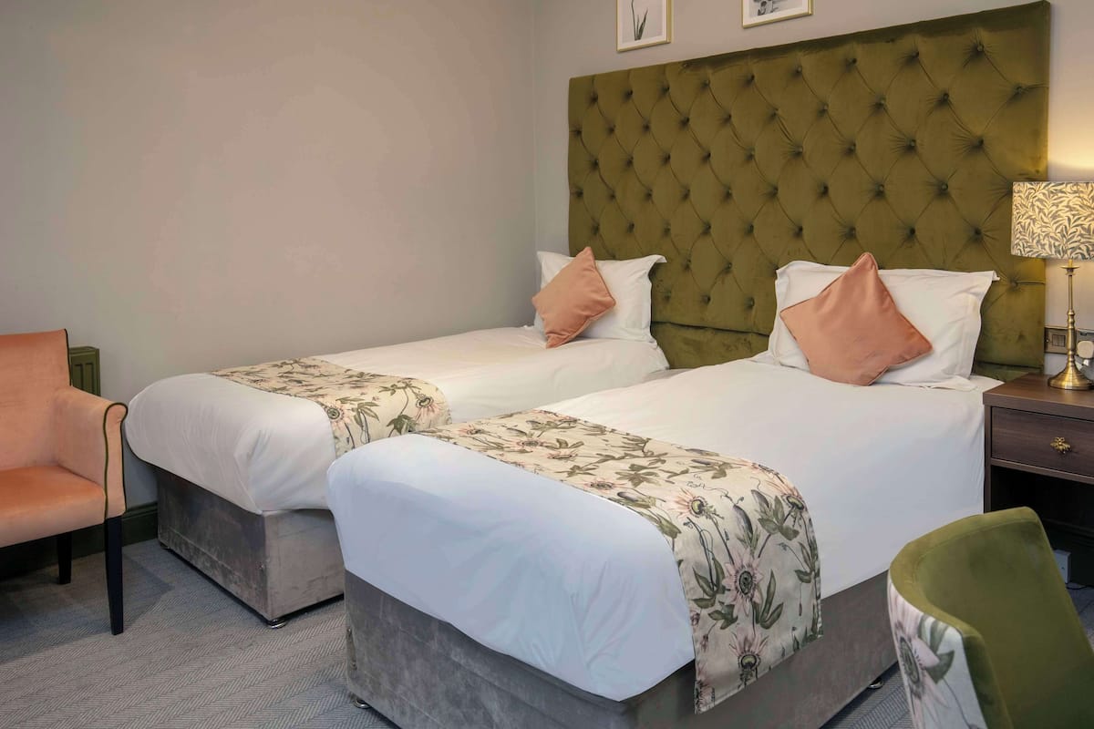 A hotel room with two beds, each with a floral bedspread and matching pillows. There is a green armchair and a green chair in the room.
