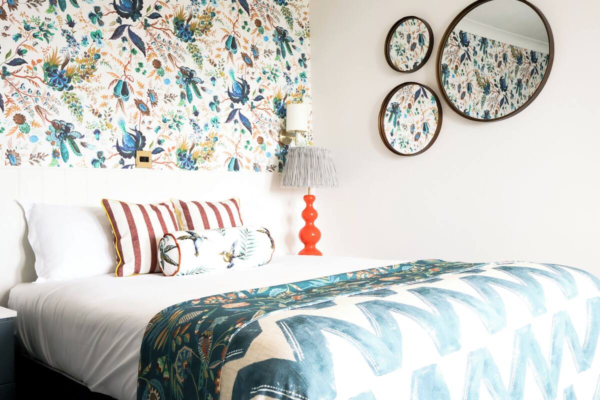 A bedroom with a floral wallpaper, a bed with patterned bedding, multiple pillows, a bedside lamp, and three wall mirrors.