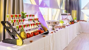 Una mesa de buffet con diversos alimentos y bebidas, incluyendo una piña y tomates, en una sala con ventanas grandes y cortinas.