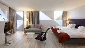 Una habitación de hotel moderna con una cama, un sofá, una silla, un televisor y ventanas amplias que dan a una vista de la ciudad.