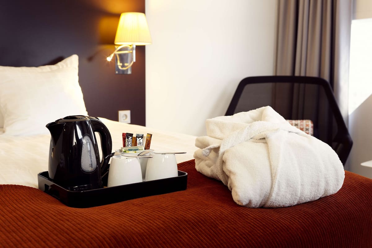 Un llit d'hotel ben fet amb una tetera negra, tasses blanques i una bata blanca sobre un edredó marró.