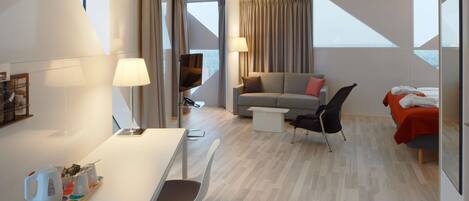 Una habitación de hotel moderna con zona de comedor, sofá, televisor y cama con una manta roja.