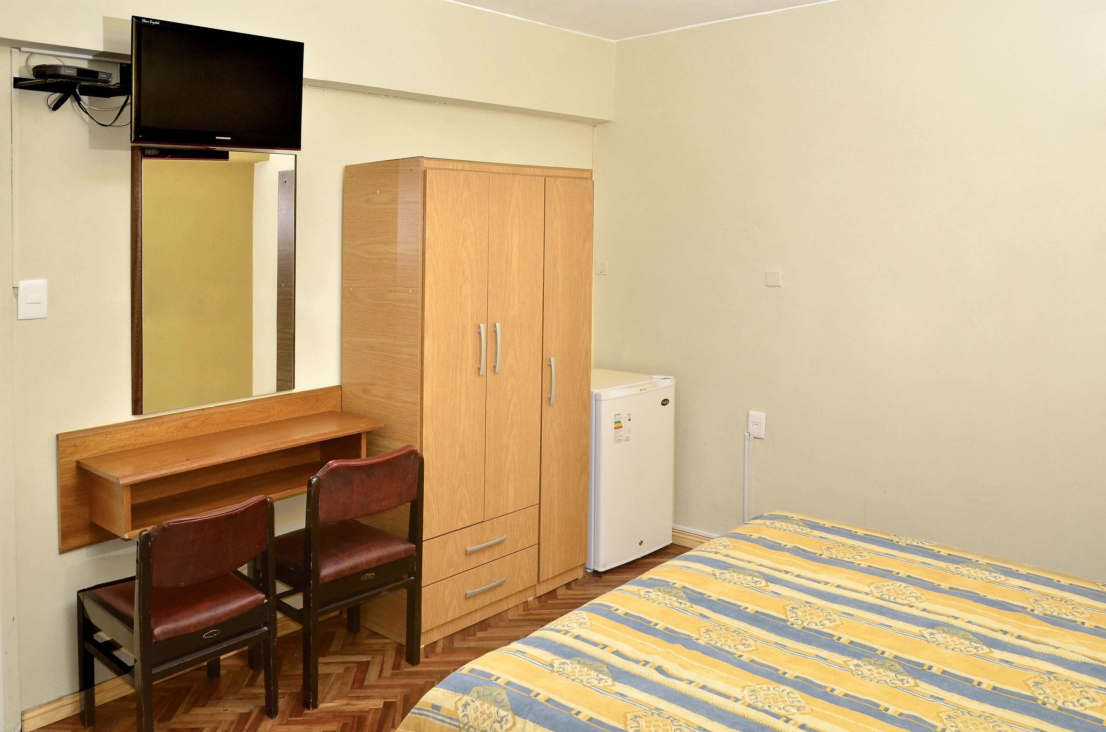 A bedroom with a bed, a wooden wardrobe, a desk with two chairs, a small white refrigerator, and a television mounted on the wall.