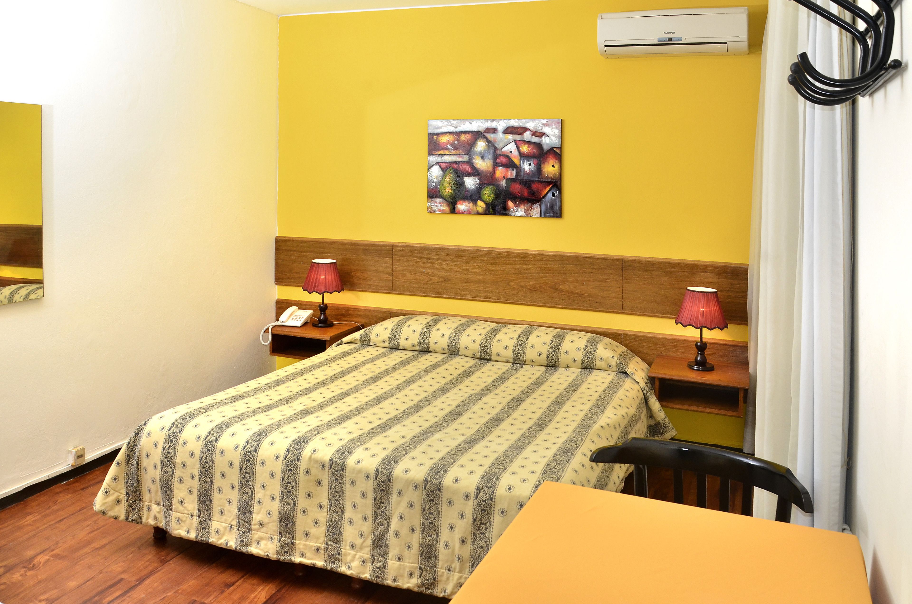 A hotel room with a bed, bedside lamps, a painting on the wall, and an air conditioning unit.