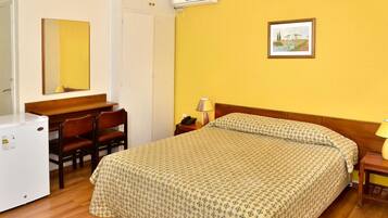 Una habitación de hotel con una cama, un escritorio con sillas, un refrigerador, una lámpara de noche y un cuadro en la pared.