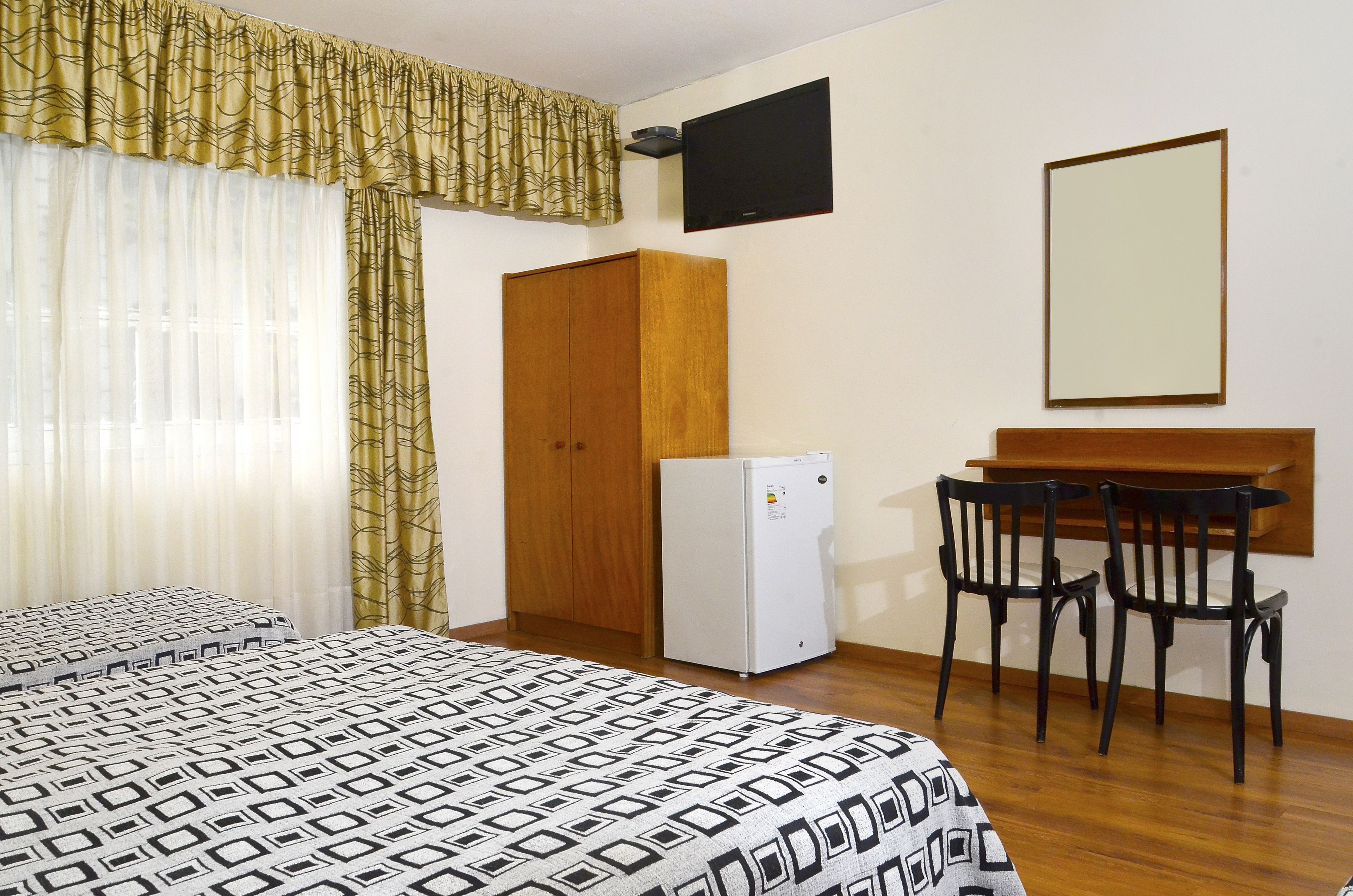 Una camera da letto con un letto, una scrivania, due sedie, un frigorifero e una televisione appesa al muro.