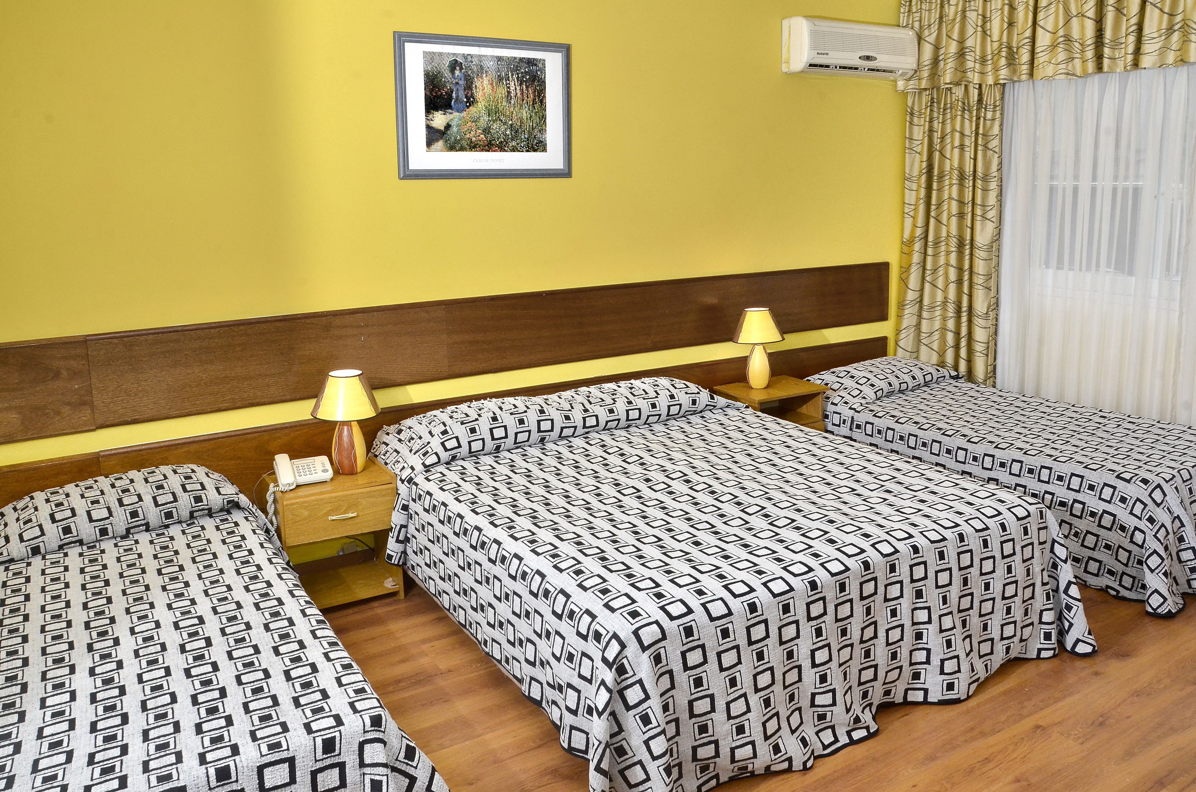 A hotel room with two single beds, a wooden headboard, a bedside table with a lamp, a framed picture on the wall, and an air conditioning unit.