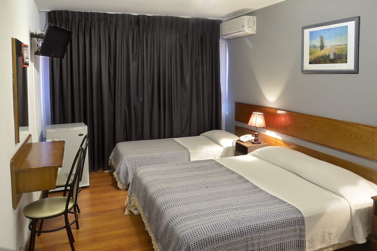 A hotel room with two beds, a desk with a chair, an air conditioner, and a framed picture on the wall.
