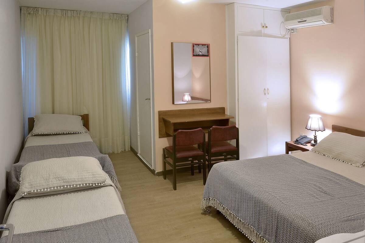 A hotel room with two single beds, a desk with two chairs, a mirror, a bedside lamp, and an air conditioning unit.