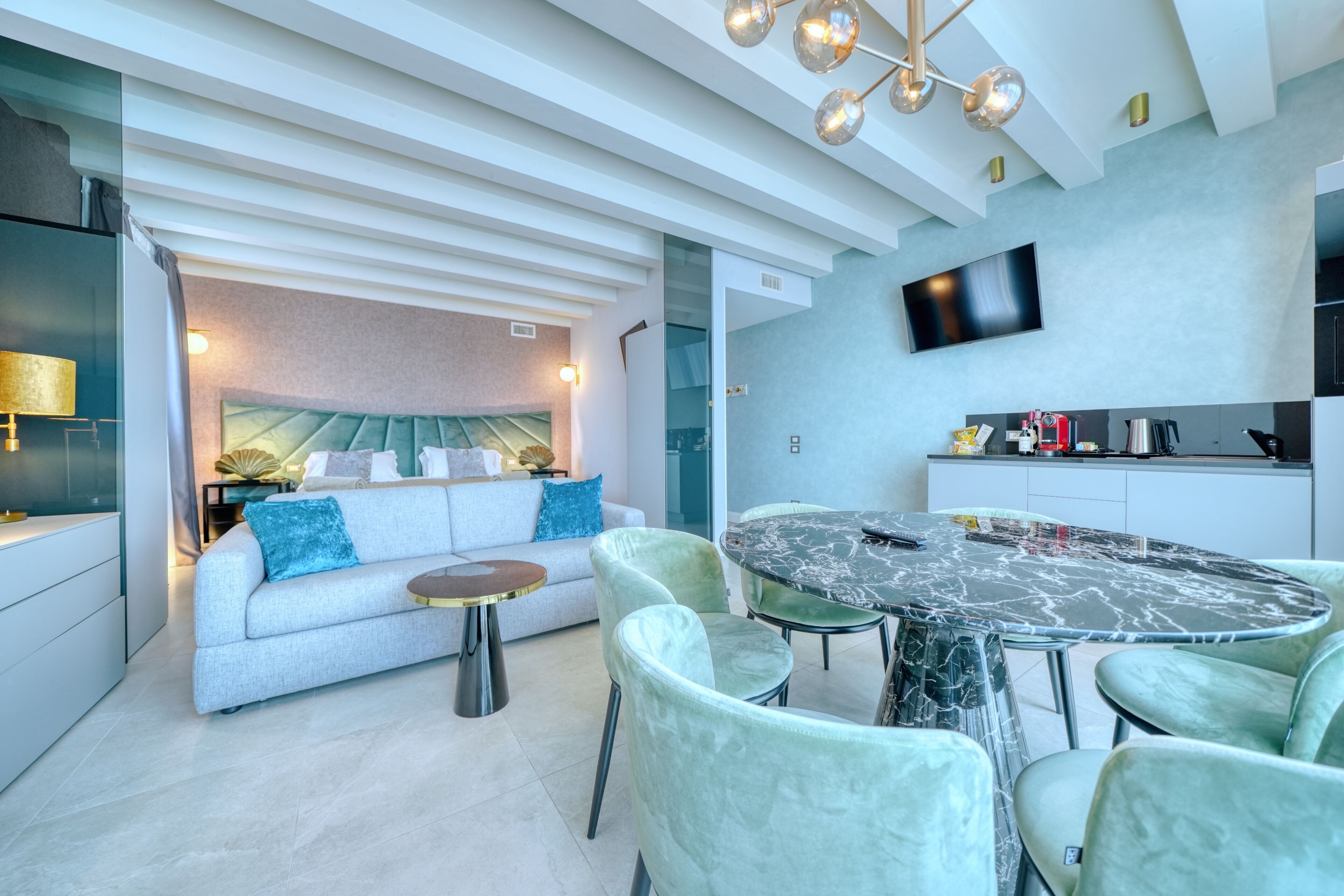 A modern living room with a sofa, a round table, and chairs, a television mounted on the wall, and a kitchen area with cabinets and appliances.
