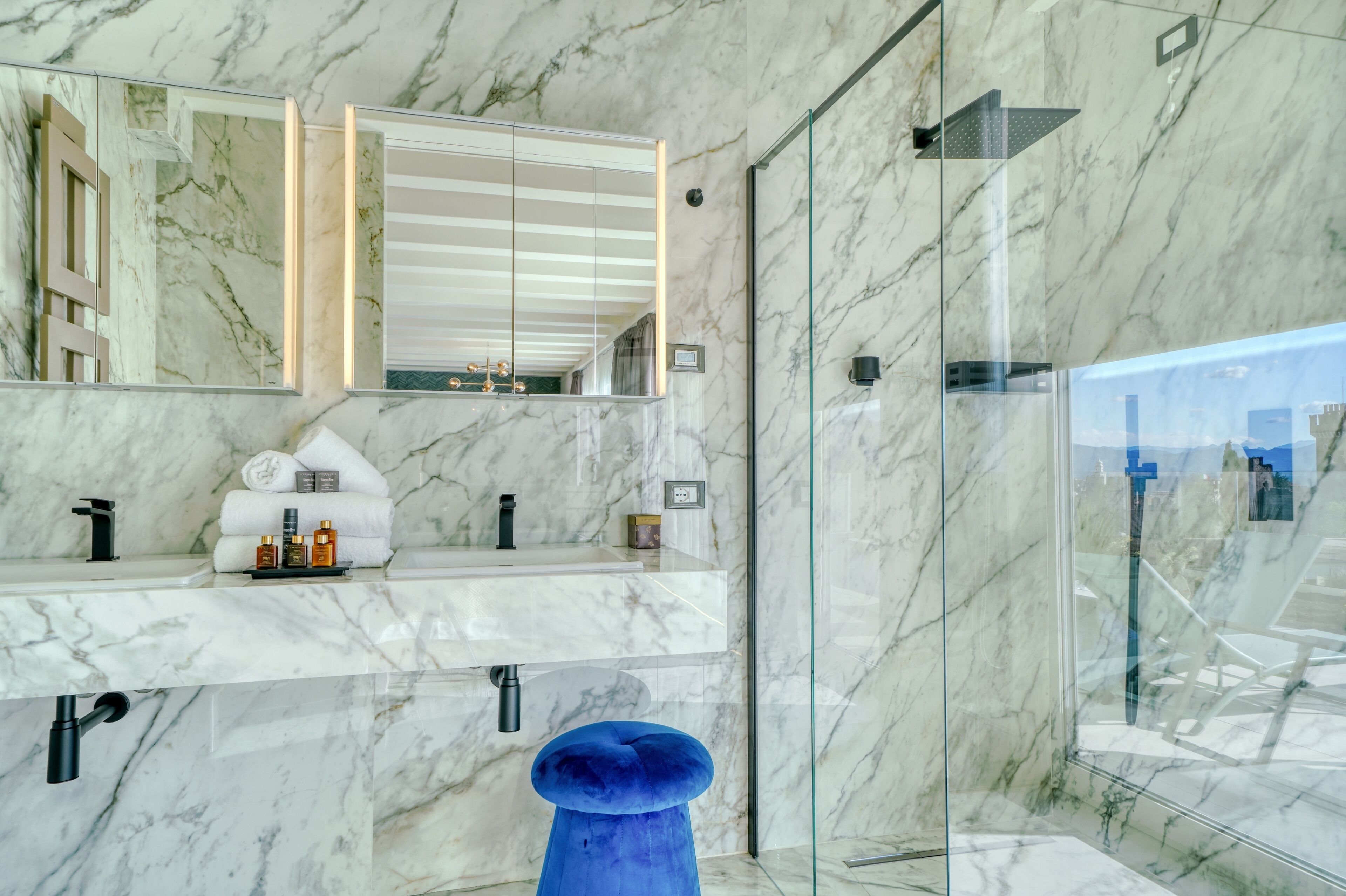Een moderne badkamer met marmeren wanden, een grote spiegel, een glazen douchecabine en een blauwe kruk.