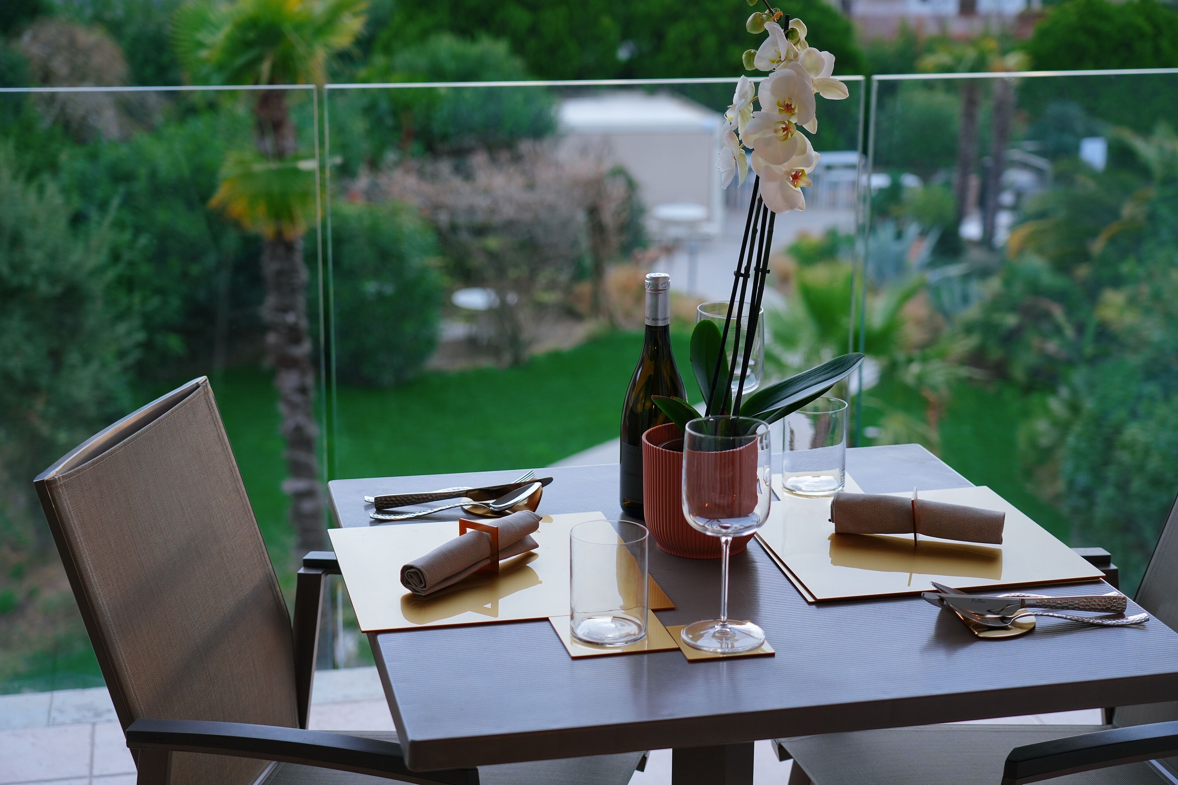 Une table à manger avec une bouteille, un verre et une orchidée, disposée sur un balcon offrant une vue sur la verdure.