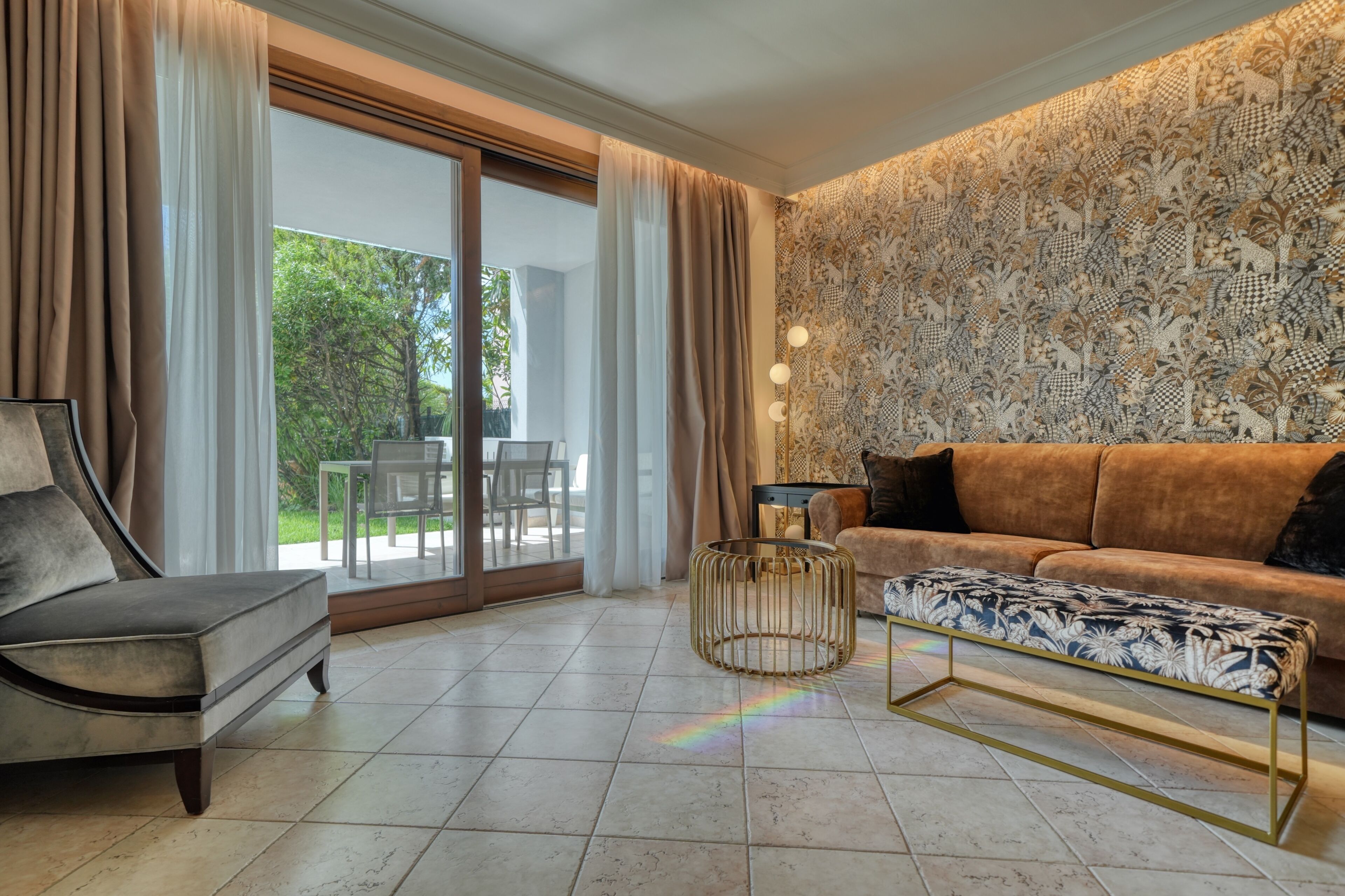 Una sala de estar con un sofá, un sillón y un reposapiés estampado. Hay una puerta de vidrio que da a un área exterior con sillas.