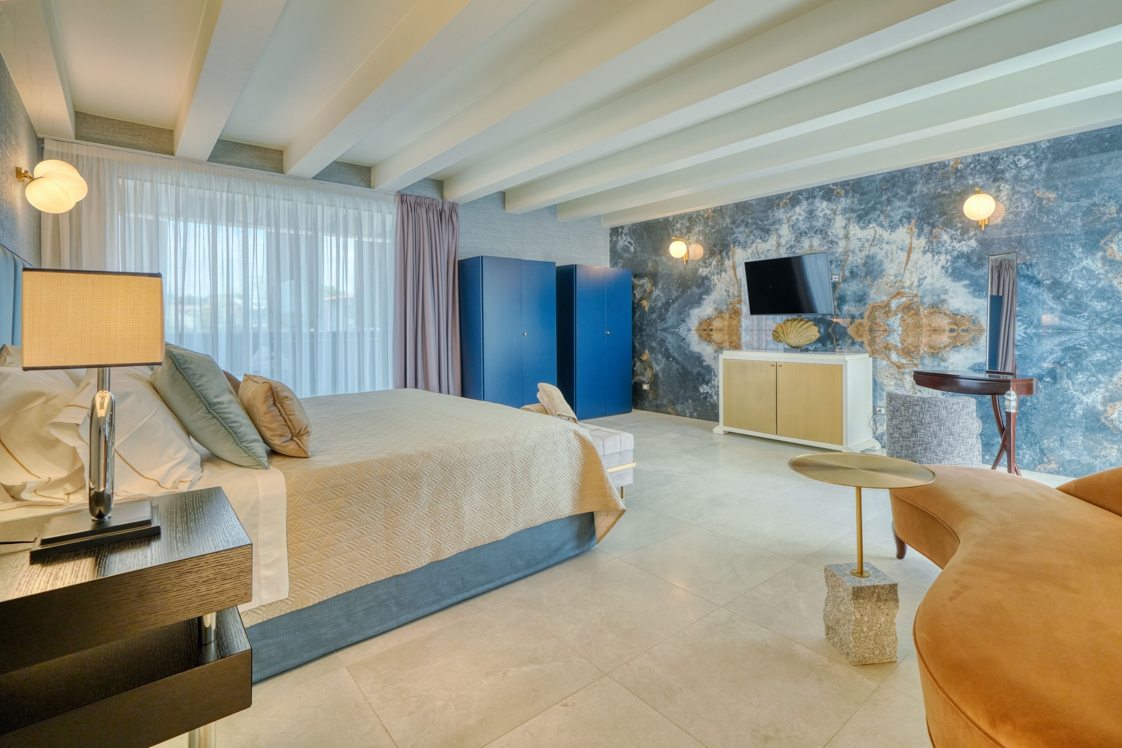 A modern hotel room with a large bed, a desk with a lamp, a TV, and a stone accent on the floor.
