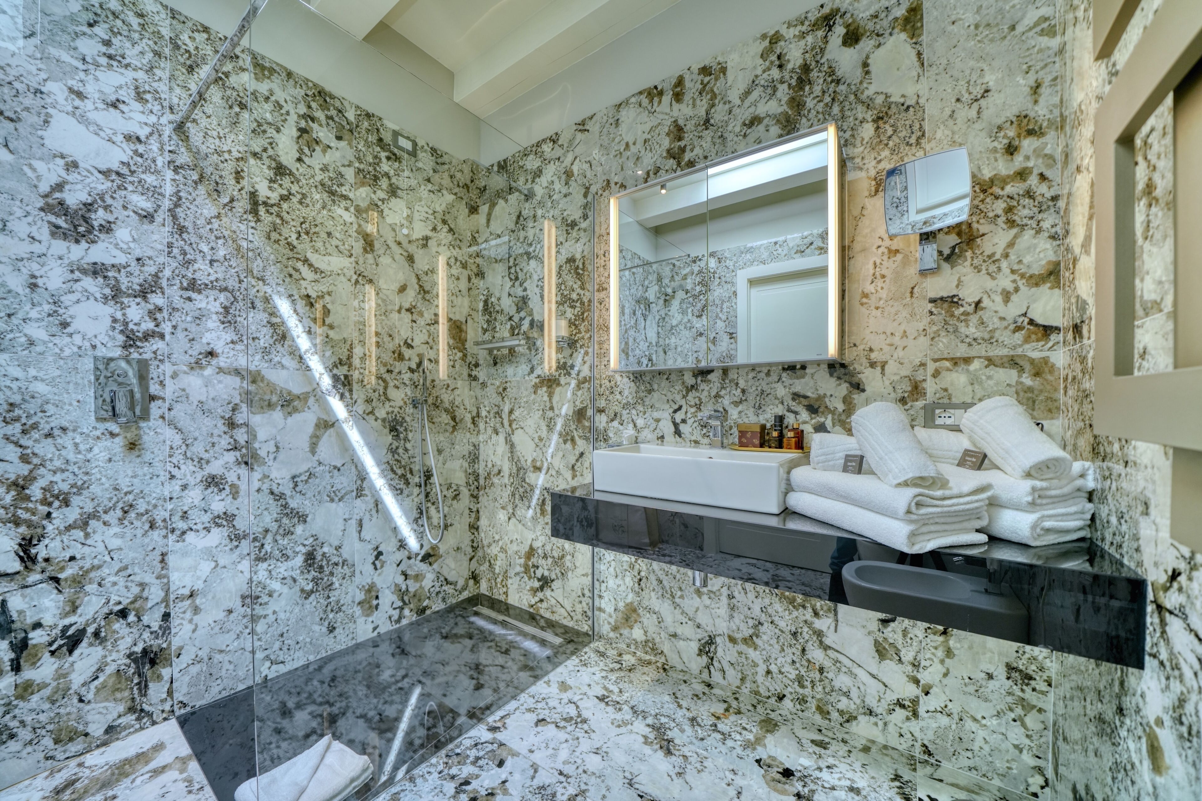 Une salle de bain avec un mur en marbre, un lavabo avec un miroir et des serviettes sur une étagère.