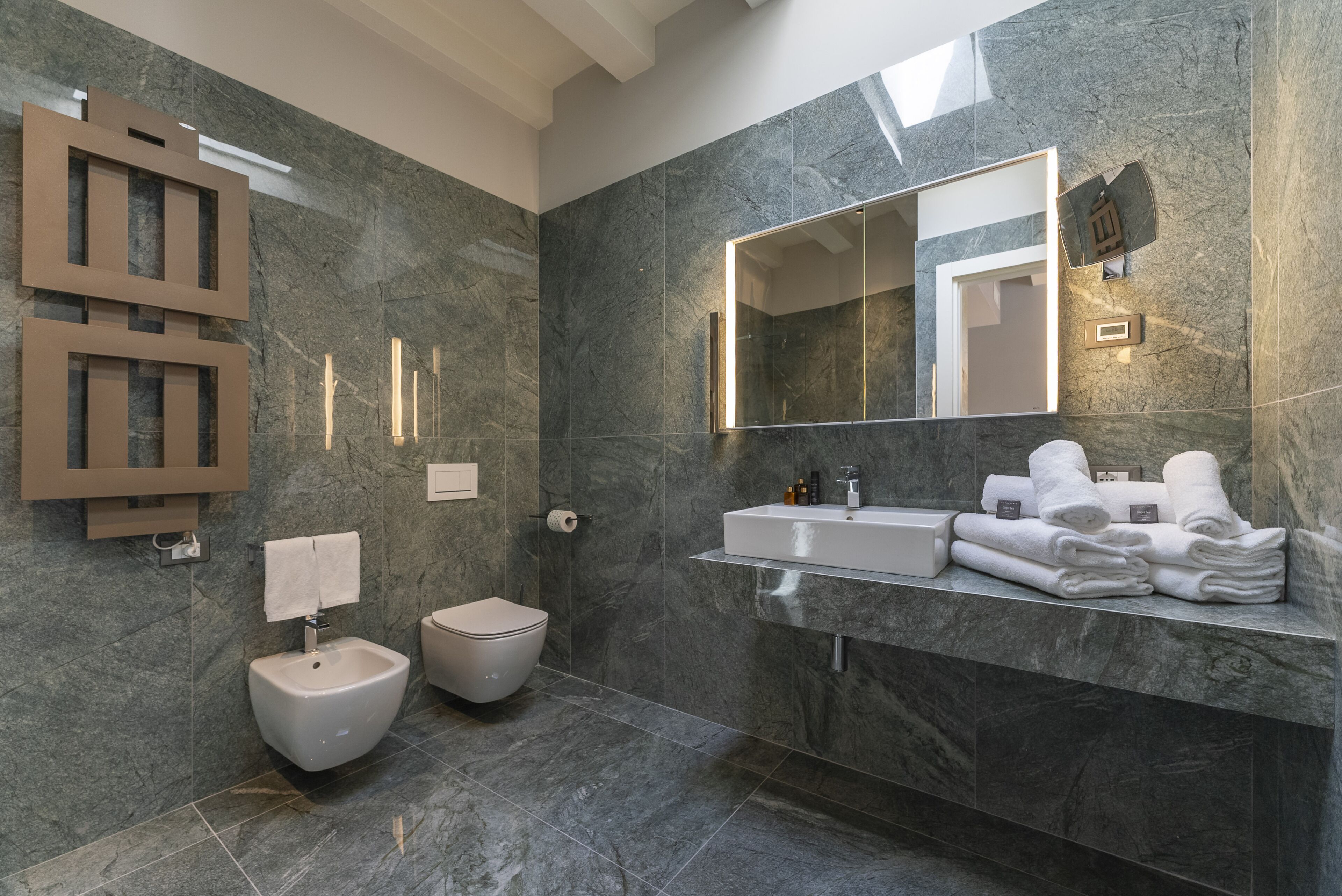 A modern bathroom with a marble wall, a large mirror, a sink, and a toilet.