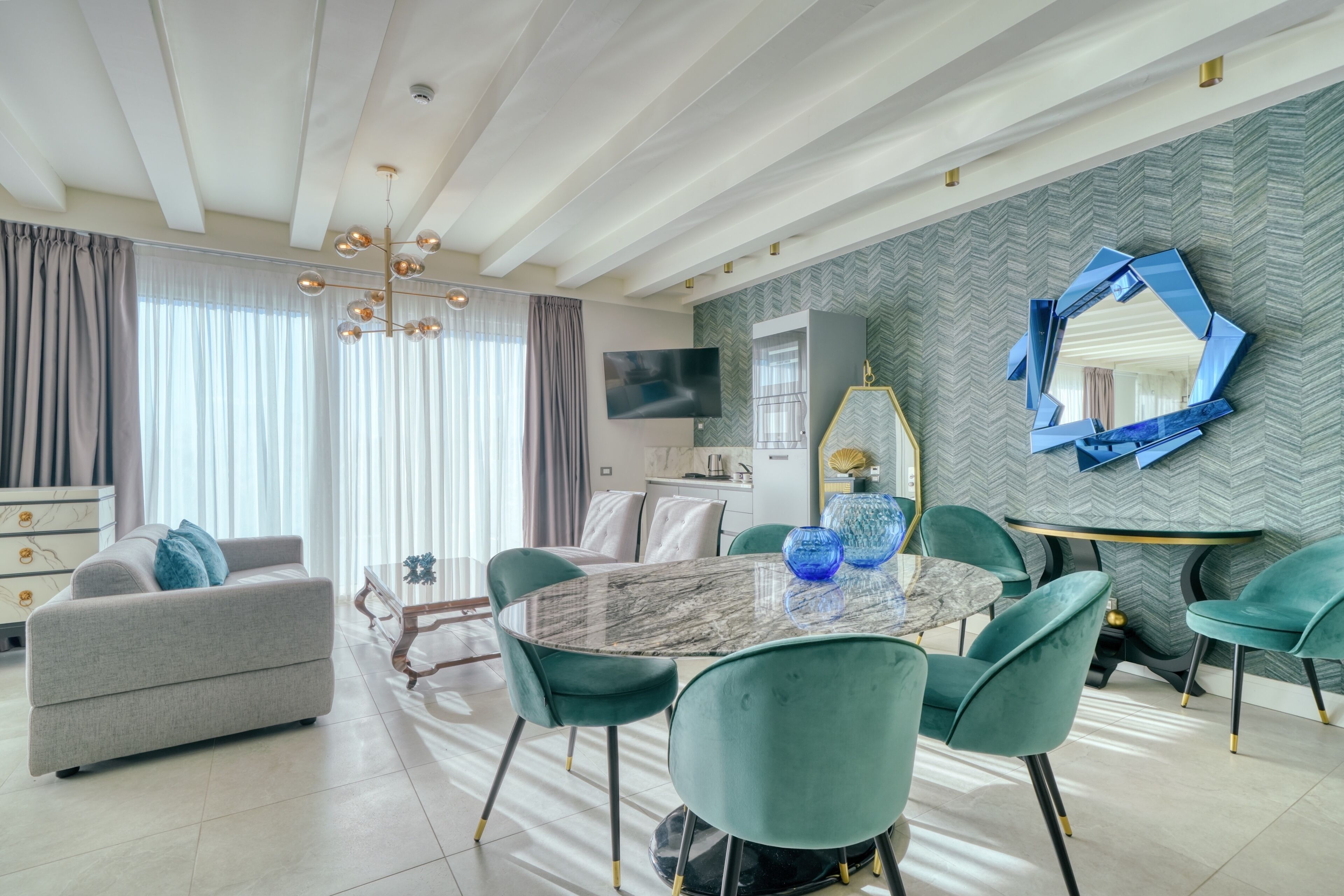 A modern living room with a marble dining table, teal chairs, a grey sofa, and a large mirror on the wall.