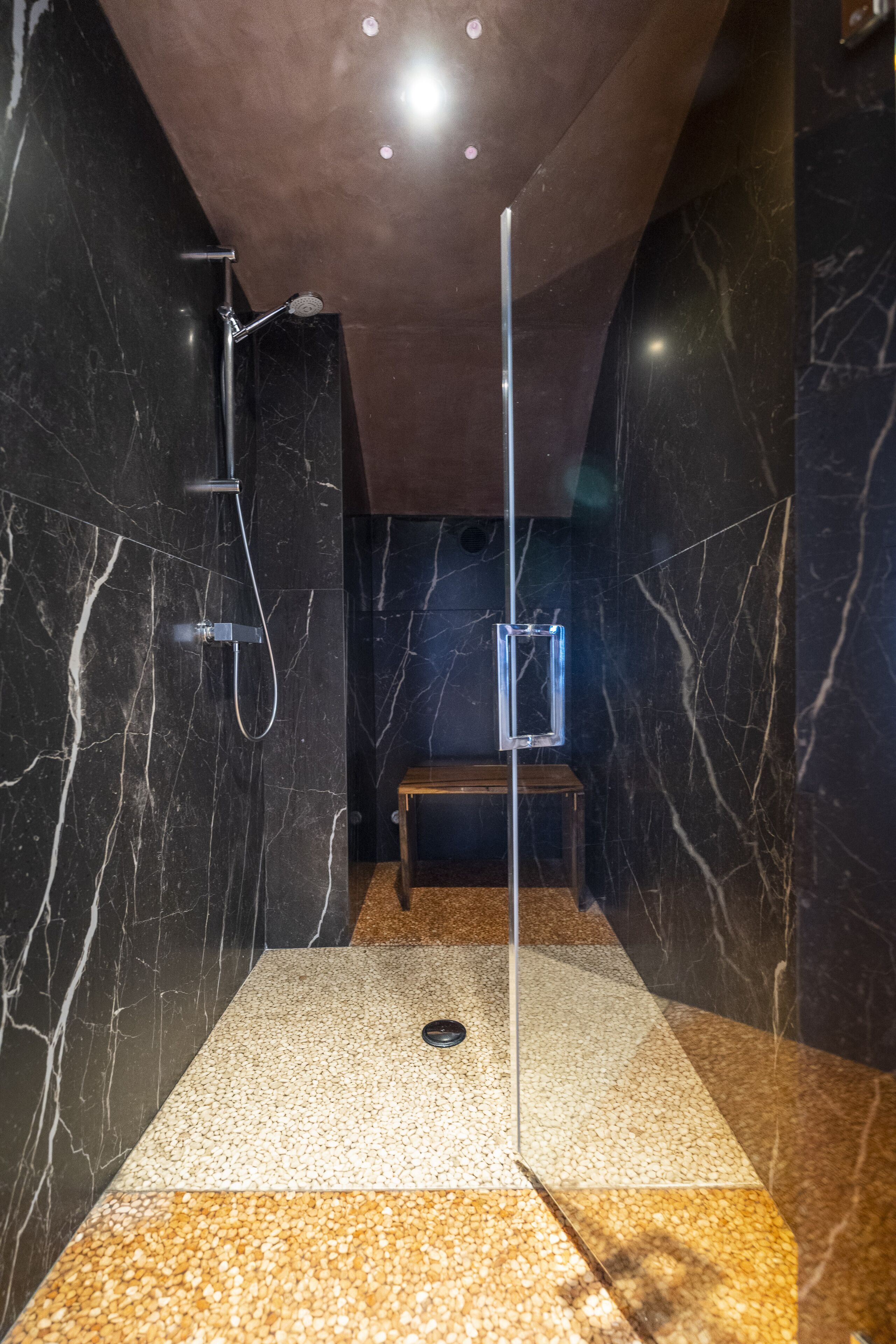 Un área de ducha moderna con mampara de vidrio, paredes de mármol y un banco.