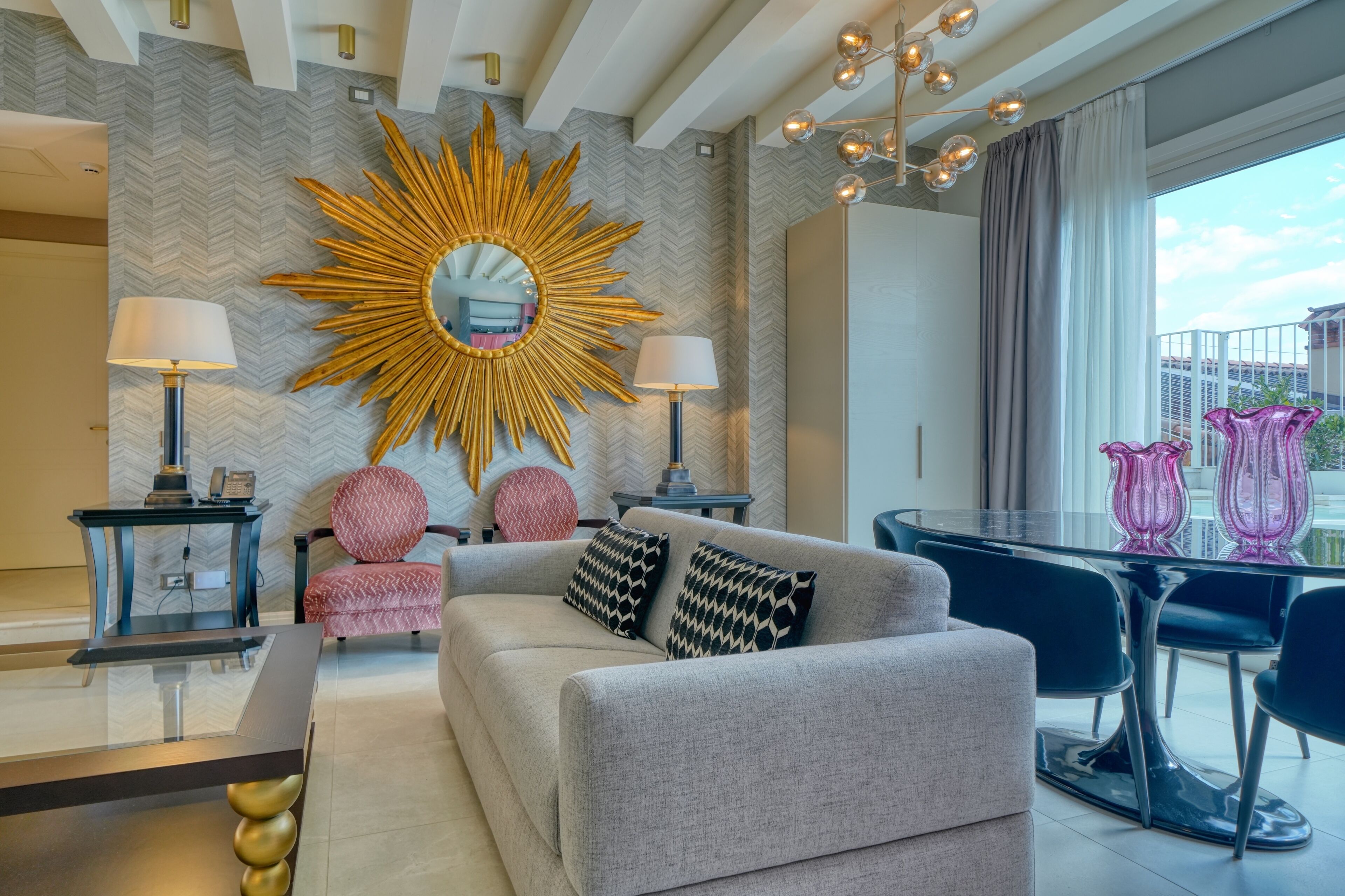 A modern living room with a sofa, two side tables, a glass coffee table, and a large mirror with a sunburst frame.