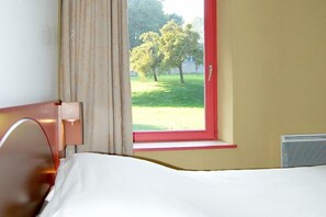 A hotel room with a bed, a window with a red frame, and a view of a grassy area with trees. - Hôtel Domaine de Volkrange (Thionville)