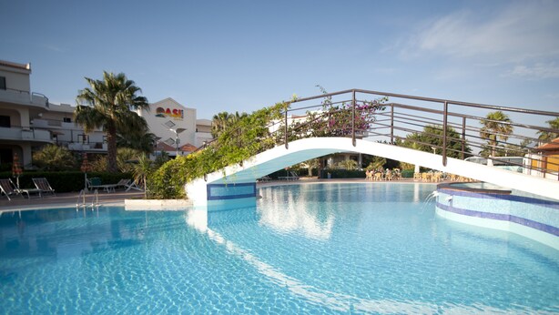 A swimming pool with a white bridge, palm trees, and a building with a sign. - Oasiclub Hotel (Vieste)