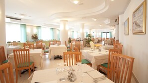 A spacious dining area with multiple tables and chairs, well-lit with ceiling lights, and large windows allowing natural light. - Oasiclub Hotel (Vieste)