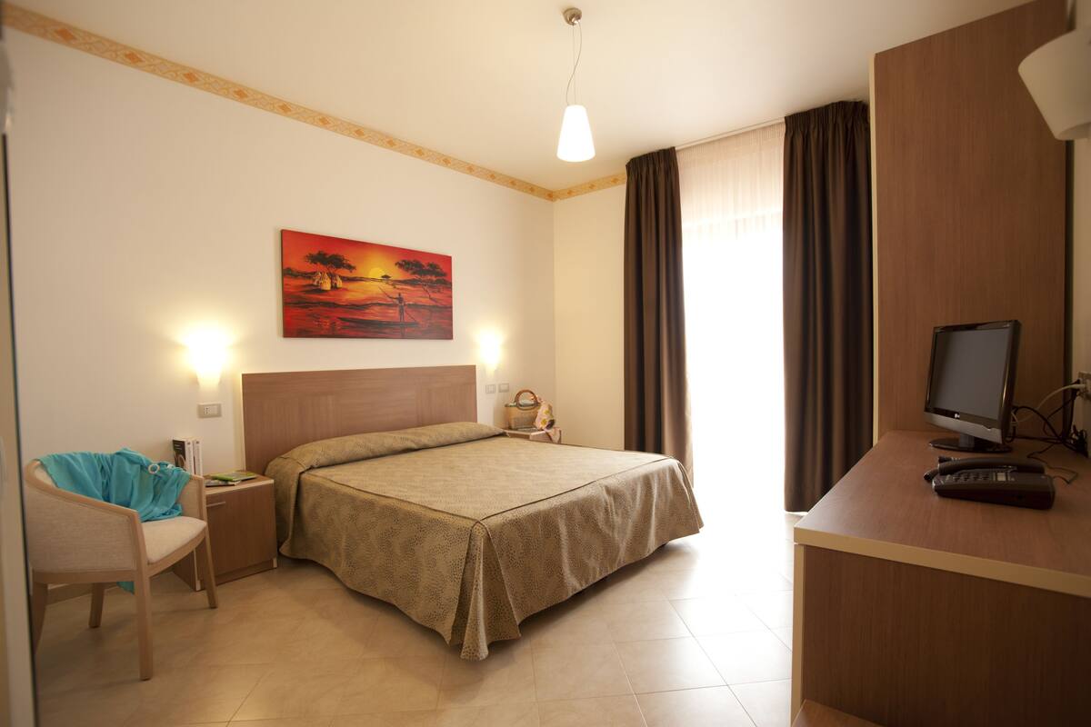 A hotel room with a bed, a desk with a computer, a chair, a nightstand, and a painting on the wall.