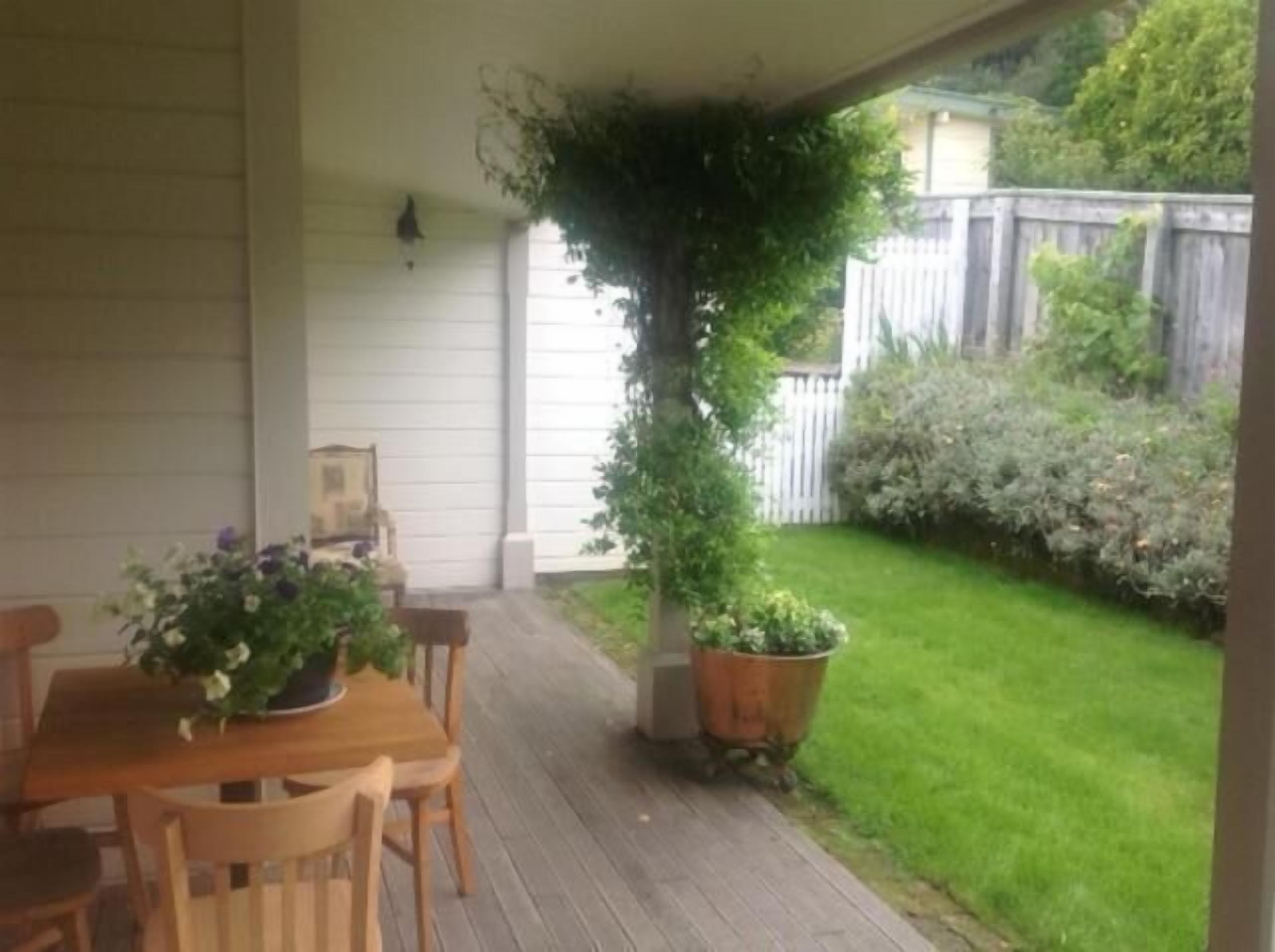 A covered patio with a wooden table and chairs, a potted plant, and a view of a grassy yard with a wooden fence.