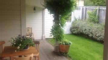 A covered patio with a wooden table and chairs, a potted plant, and a view of a grassy yard with a wooden fence.