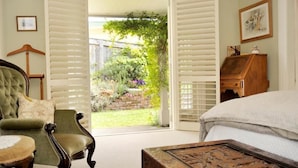 Une chambre à coucher avec un lit, un fauteuil vert, une commode en bois et une vue sur un jardin, visible à travers une porte dotée de volets blancs.