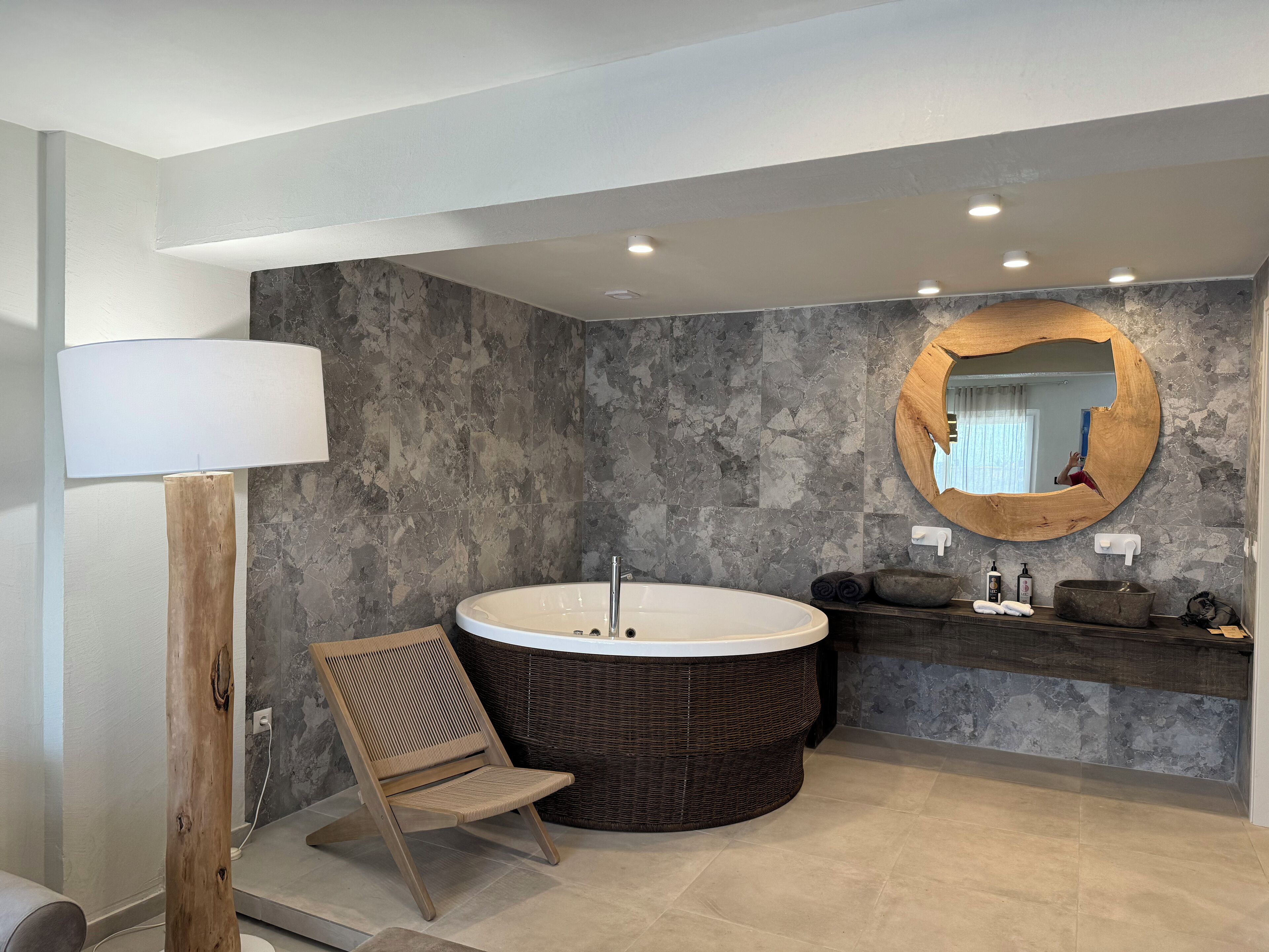 A modern bathroom with a freestanding bathtub, a wooden chair, a large mirror, and a stone-textured wall.